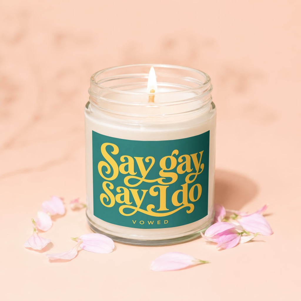 Candle with lit wick and teal label reading "Say gay Say I do" below flowers.