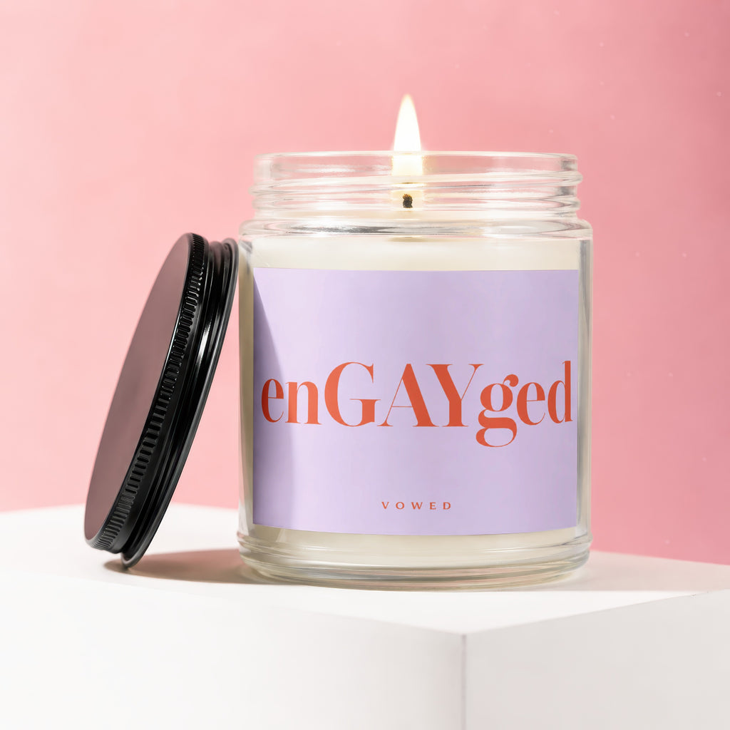 Glass jar candle with "enGAYged" and "VOWED" on a lavender label, lit wick.