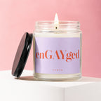 Glass jar candle with "enGAYged" and "VOWED" on a lavender label, lit wick.