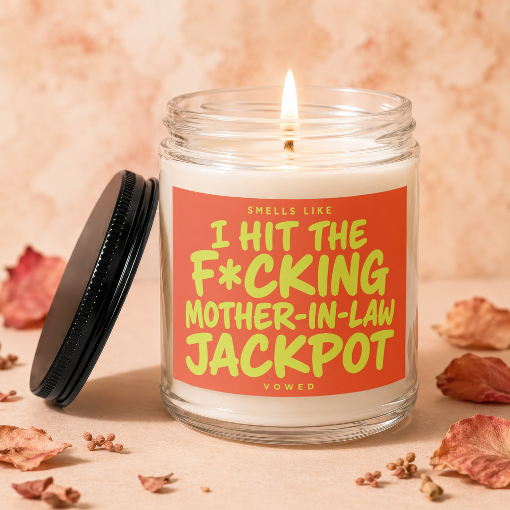 Glass candle with lit wick and black lid, reading "Smells like I hit the f*cking mother-in-law jackpot."