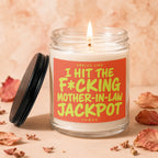 Glass candle with lit wick and black lid, reading "Smells like I hit the f*cking mother-in-law jackpot."