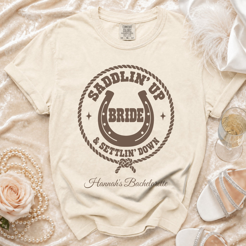 Cream-colored t-shirt featuring a brown western-themed design that reads Saddlin Up Bride and Settlin Down with a horseshoe.