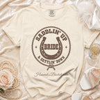 Cream-colored t-shirt featuring a brown western-themed design that reads Saddlin Up Bride and Settlin Down with a horseshoe.
