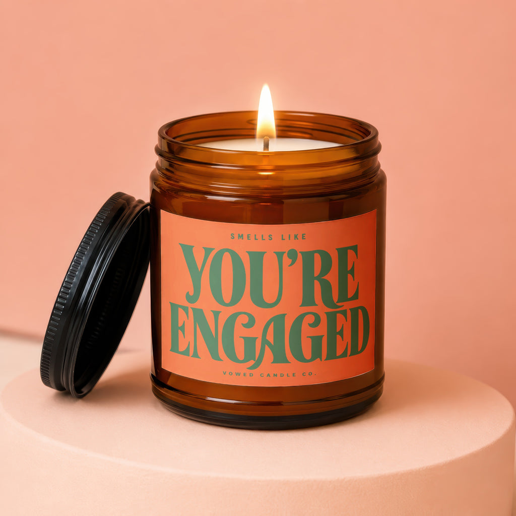 Amber candle with lit wick and orange label that reads "Smells Like You're Engaged".