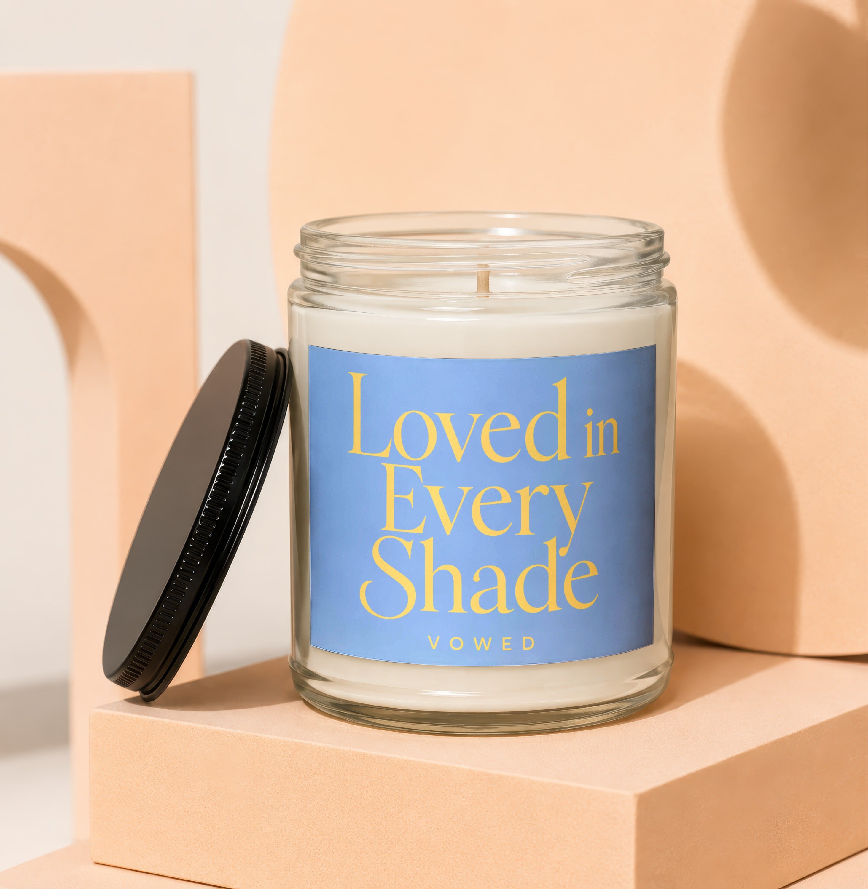 Candle with "Loved in Every Shade" text on a blue label, black lid nearby.