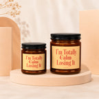 Two amber glass candles with black lids, featuring yellow labels that read "I'm Totally Calm Losing It."