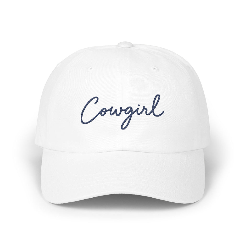 A white baseball cap with the word Cowgirl embroidered in blue cursive on the front panel.
