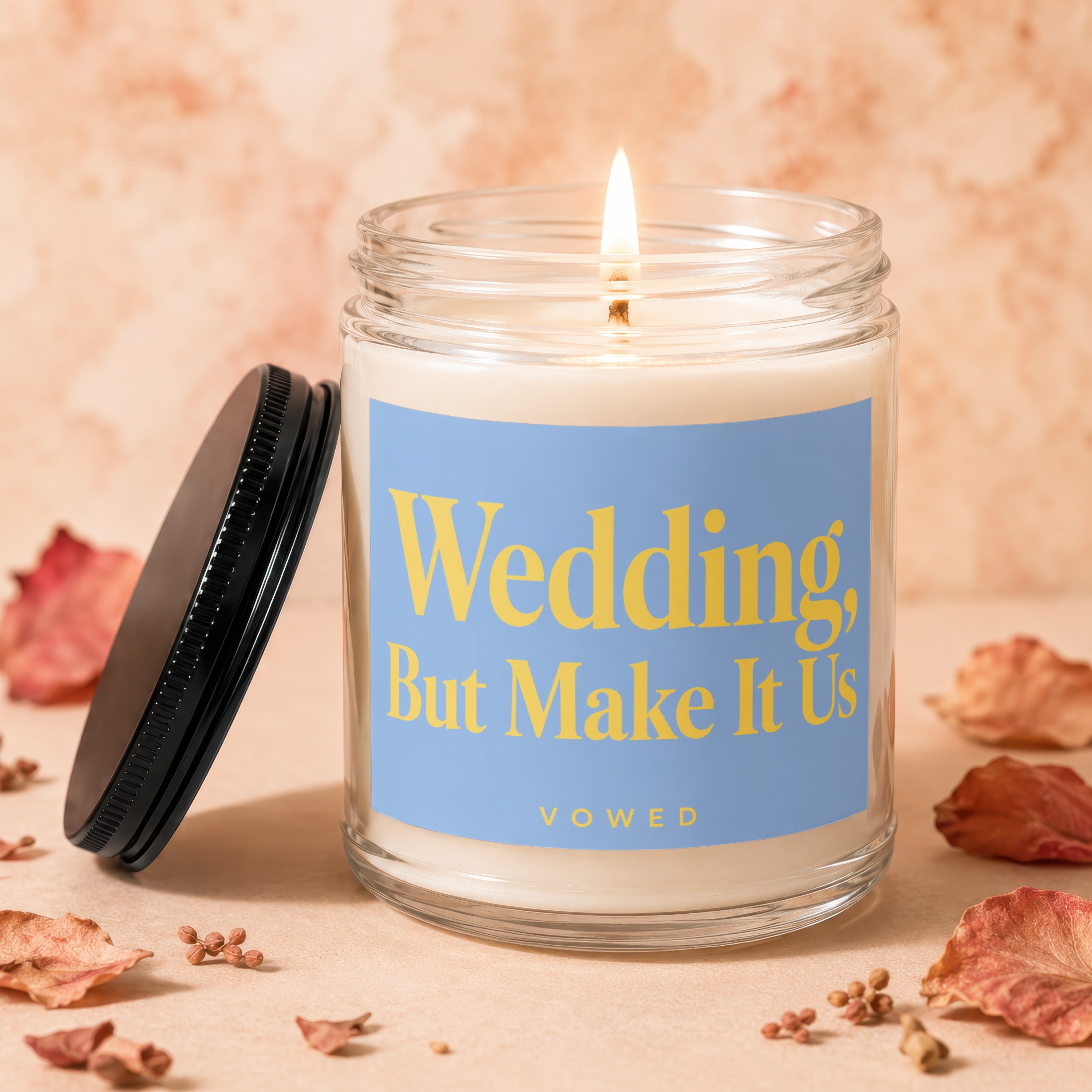 Glass jar candle with lit flame, reading "Wedding, But Make It Us".