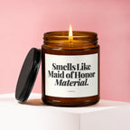 Brown glass candle with lit wick, lid off, reading "Smells Like Maid of Honor Material."