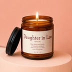 Amber glass candle with burning wick, lid off, reads "Daughter in Law (NOUN)" definition.