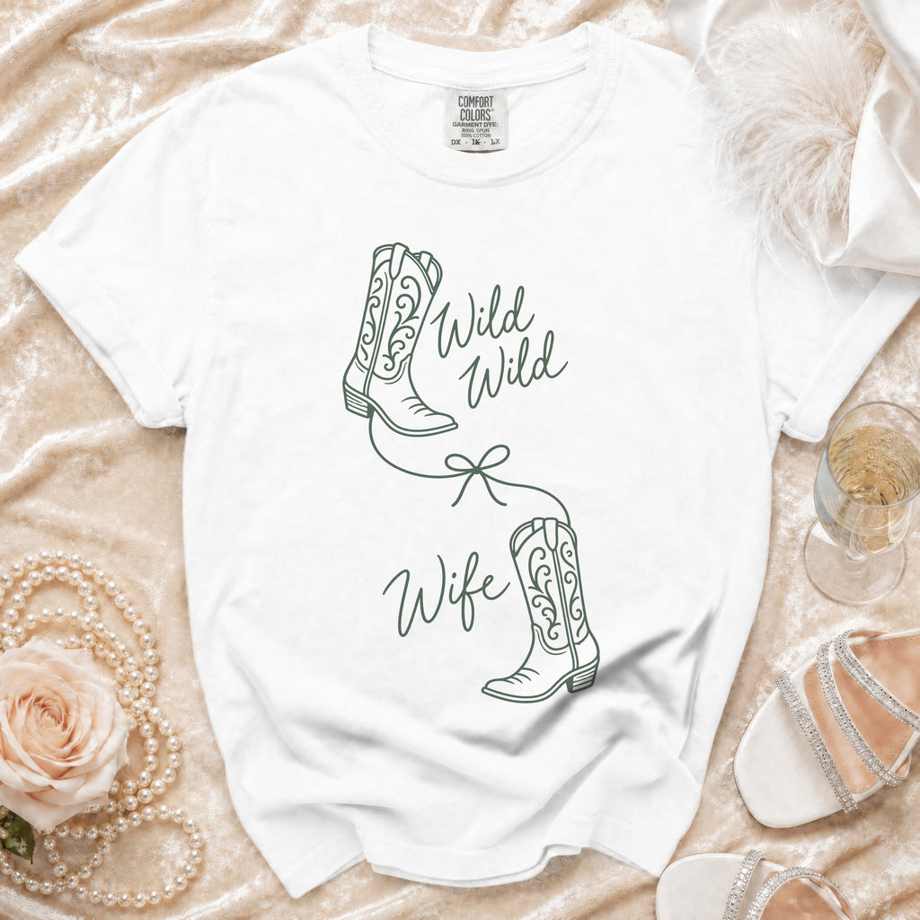 A white crewneck t-shirt features a green line-art graphic of two cowboy boots tied with a bow, with the text "Wild Wild Wife".