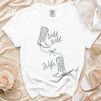 A white crewneck t-shirt features a green line-art graphic of two cowboy boots tied with a bow, with the text "Wild Wild Wife".