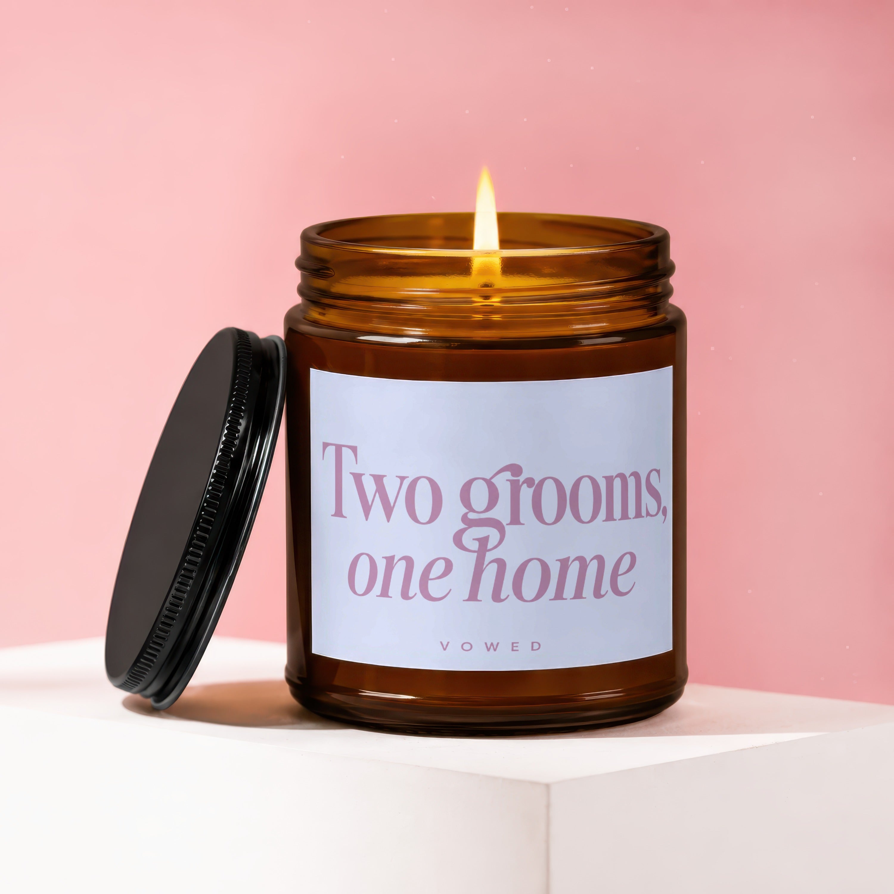 Amber glass candle with lit wick, black lid, and label reading "Two grooms, one home VOWED".