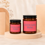 Two amber glass candles with pink labels reading "GOOD LUCK, Babe." and "VOWED".