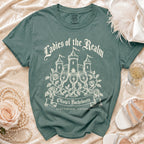 A slate green t-shirt features a castle graphic, wine glasses, roses, and the text "Ladies of the Realm" and "Olivia's Bachelorette Scottsdale, Arizona".