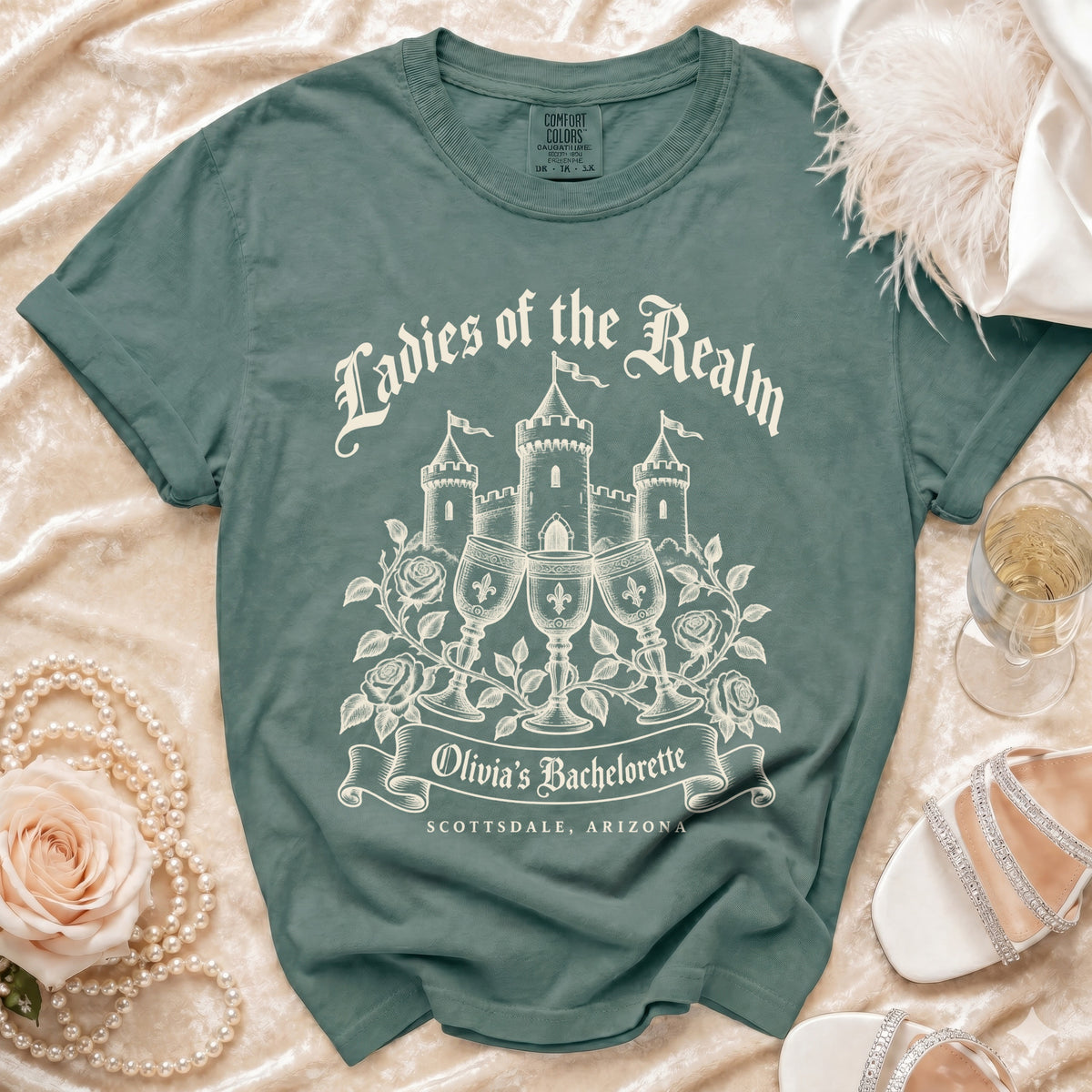 A slate green t-shirt features a castle graphic, wine glasses, roses, and the text "Ladies of the Realm" and "Olivia's Bachelorette Scottsdale, Arizona".