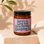 Amber jar candle with pink label reading "Here's to Love and Early Mornings Sleeping In".