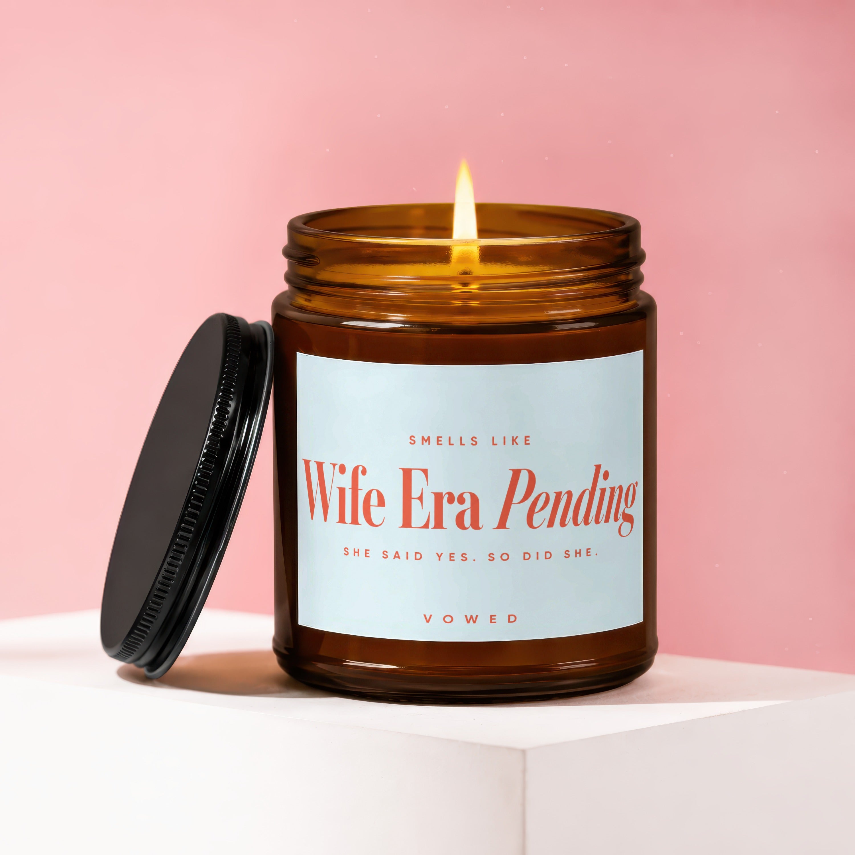 Amber jar candle with label reading "Smells like Wife Era Pending" on pink background.