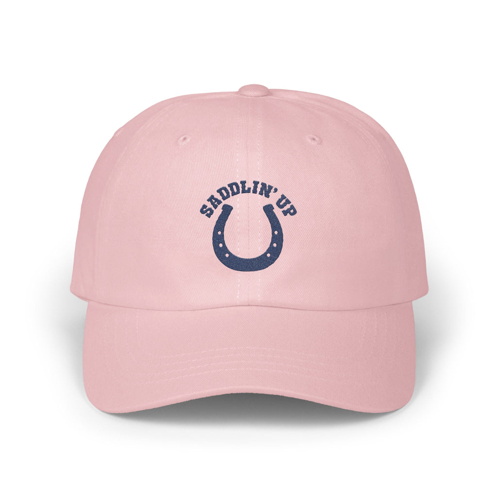 A pink baseball cap featuring a navy blue embroidered horseshoe and the text SADDLIN' UP.