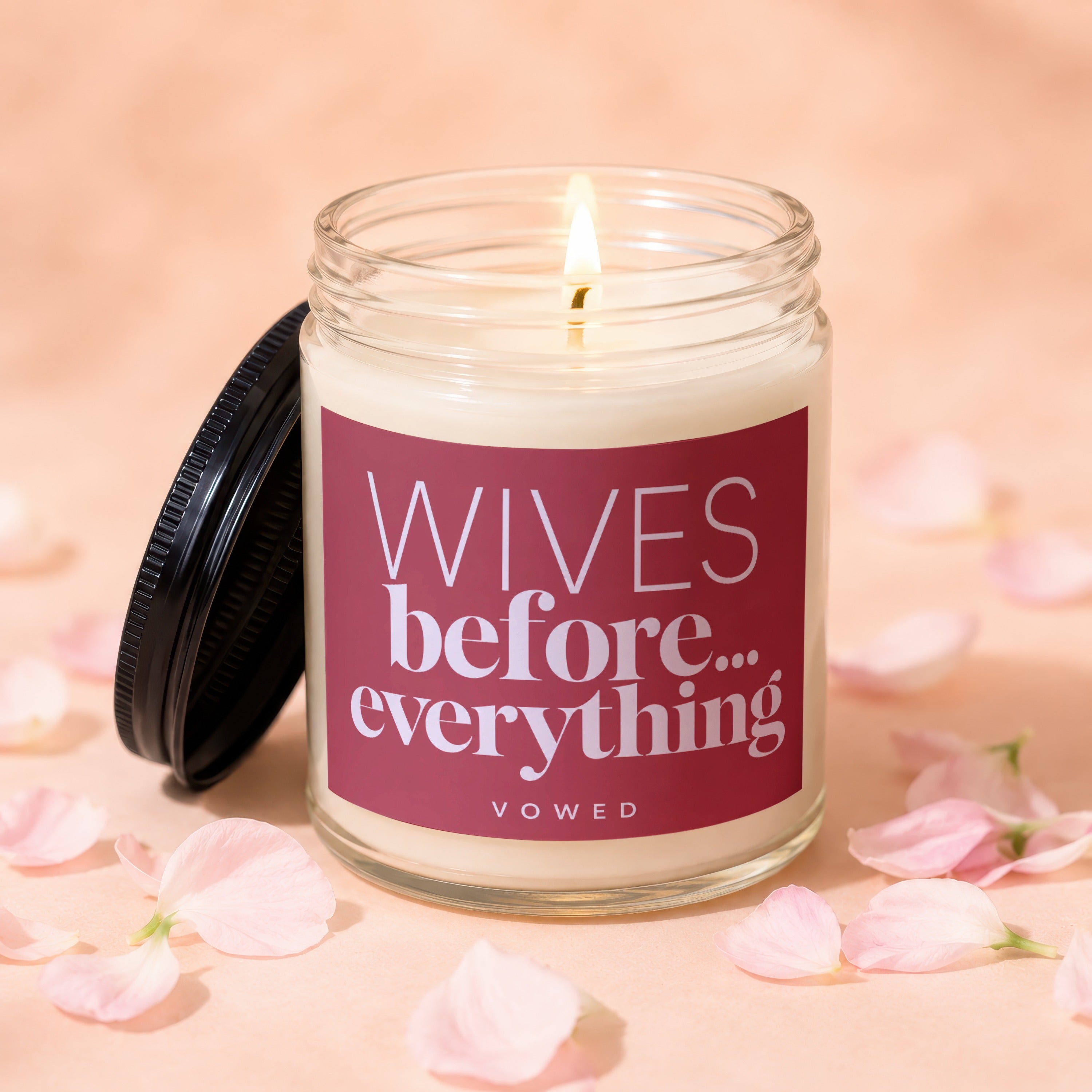 Glass candle with lit wick, lid off, reads "Wives before... everything"