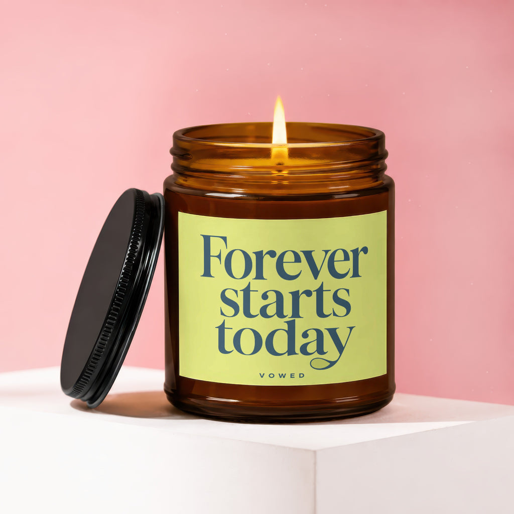 Amber glass candle with lit flame, lid ajar, features text "Forever starts today Vowed".