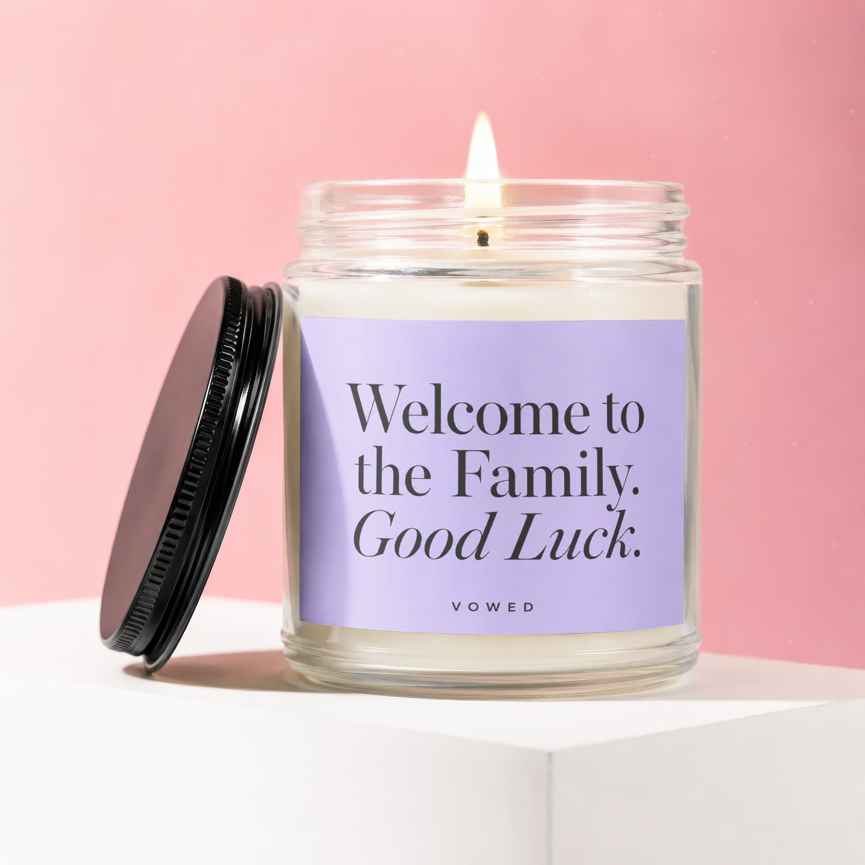 Candle with a lit wick on a light surface, violet label says "Welcome to the Family. Good Luck."