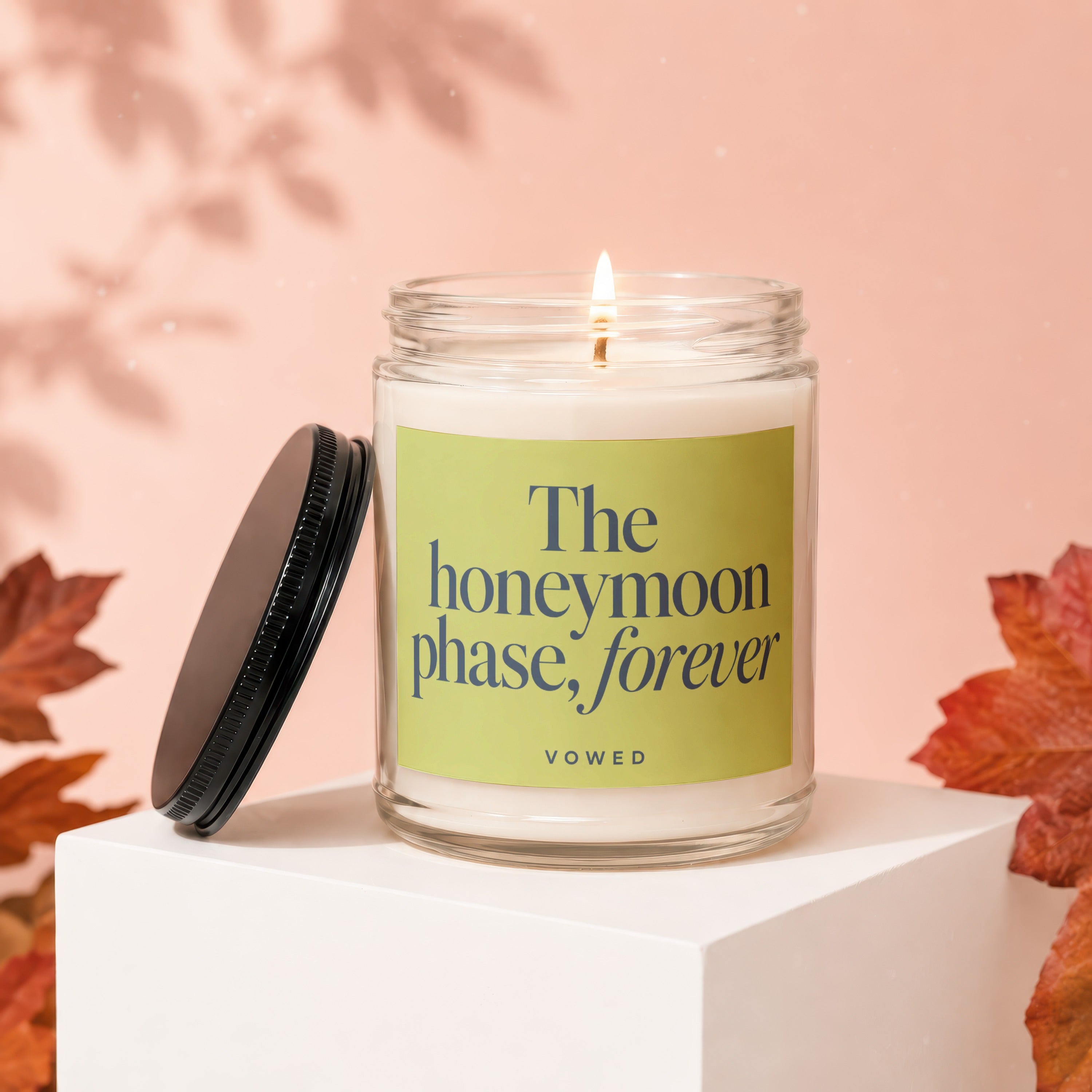 Lit candle in a glass jar with a black lid, displaying "The honeymoon phase, forever" text.