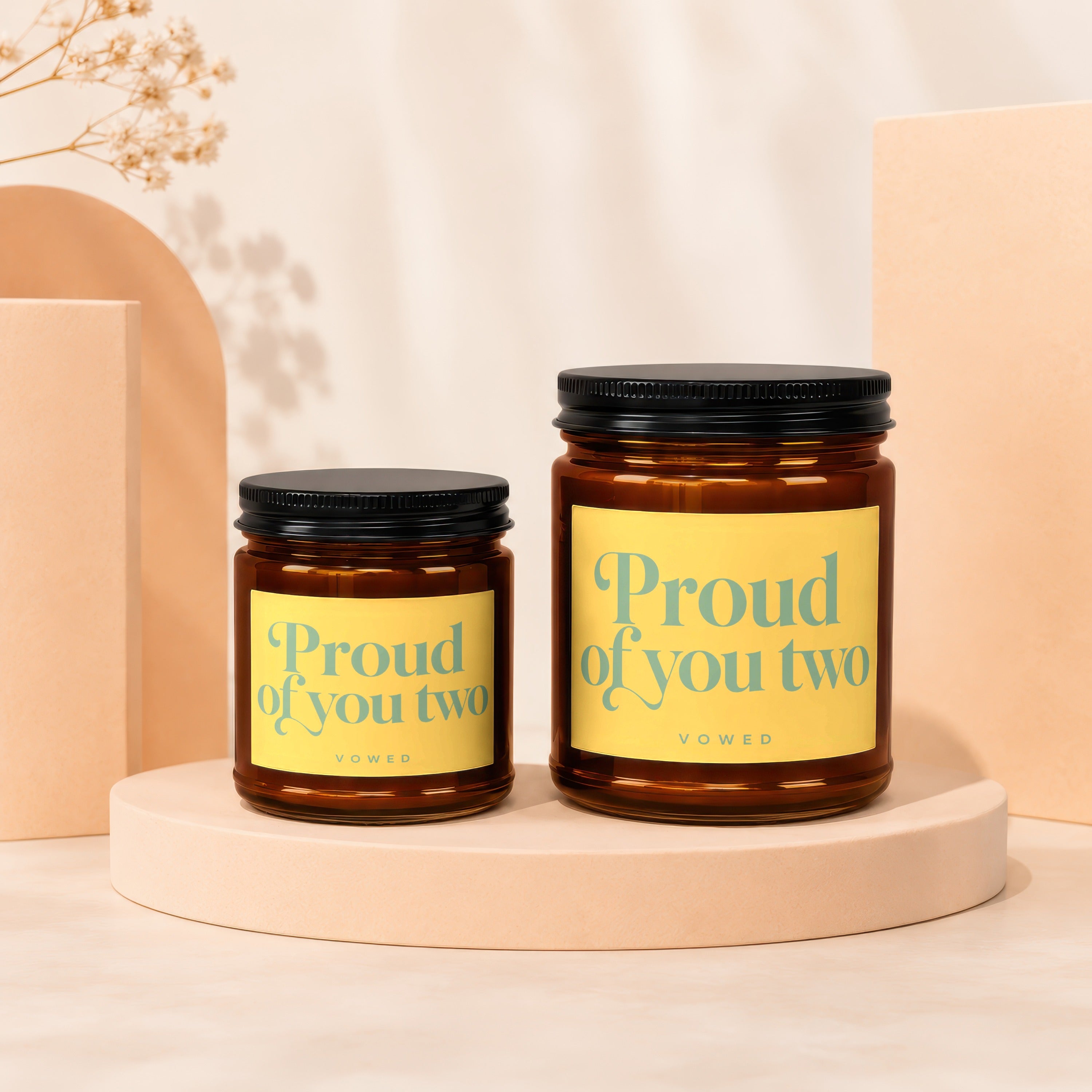 Two amber glass candles with black lids and yellow labels reading "Proud of you two".