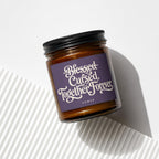 Amber jar candle with "Blessed Cursed Together Forever" text and "vowed" on purple label.