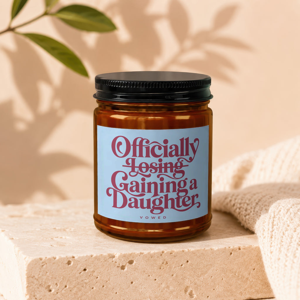 Amber jar candle with a blue label reading "Officially Losing Gaining a Daughter."