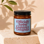 Amber jar candle with a blue label reading "Officially Losing Gaining a Daughter."