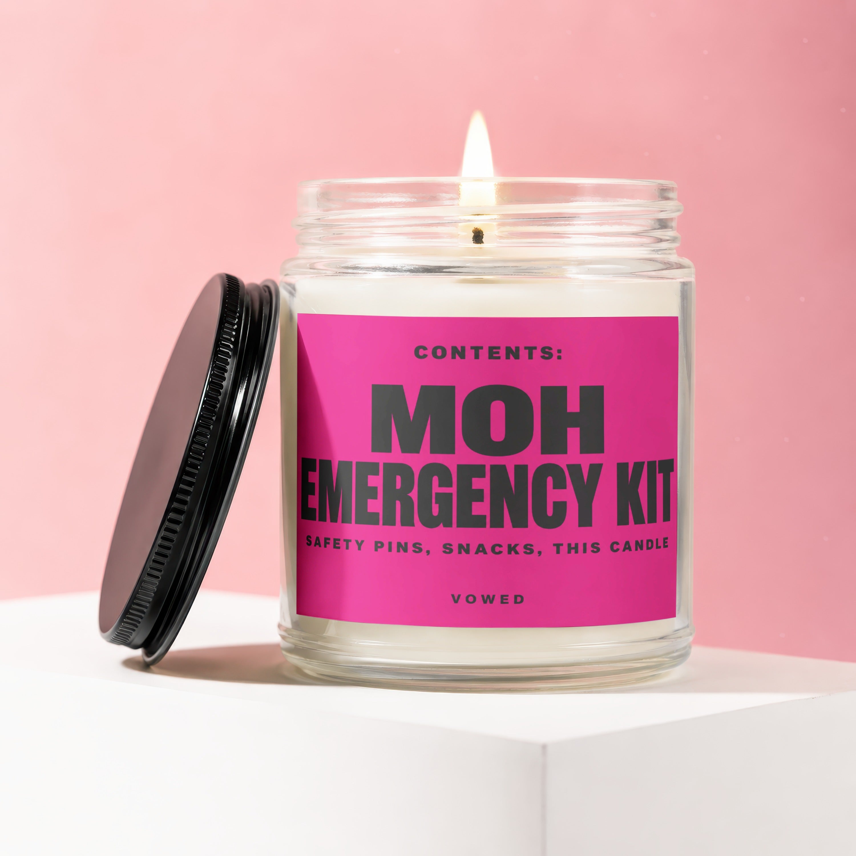 Burning candle in jar labeled "MOH EMERGENCY KIT" and "VOWED" with pink background.