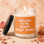 Glass jar candle with lit wick and orange label reading "Welcome to My Dream Budget Wedding"