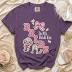 Purple t-shirt with "Bride In My Bride Era" graphic, pink bow, flowers, and champagne glasses.
