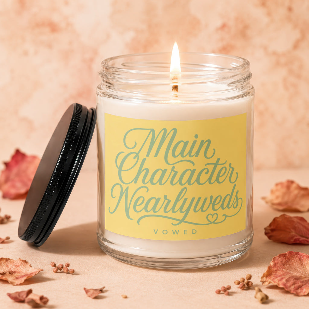 Glass jar candle with lit wick, yellow label reads "Main Character Nearlyweds"