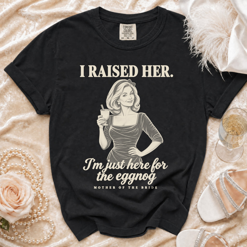 A black t-shirt printed with retro-style graphics, holding a glass and text reading I raised her. I am just here for the eggnog. Mother of the bride.