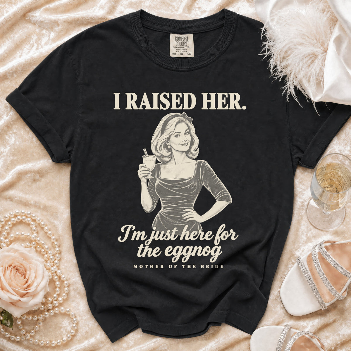 A black t-shirt printed with retro-style graphics, holding a glass and text reading I raised her. I am just here for the eggnog. Mother of the bride.