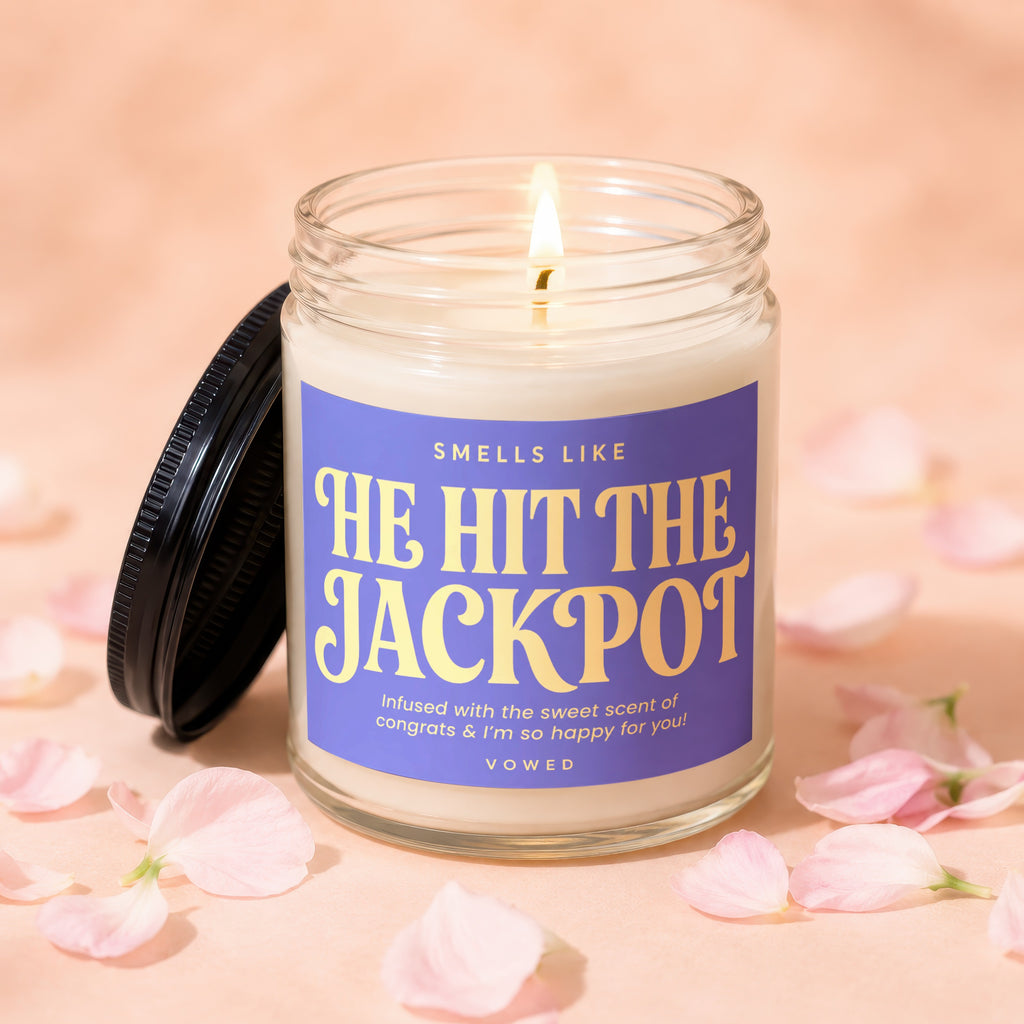 Vowed candle labeled "He Hit The Jackpot" with lit wick amidst pink petals.