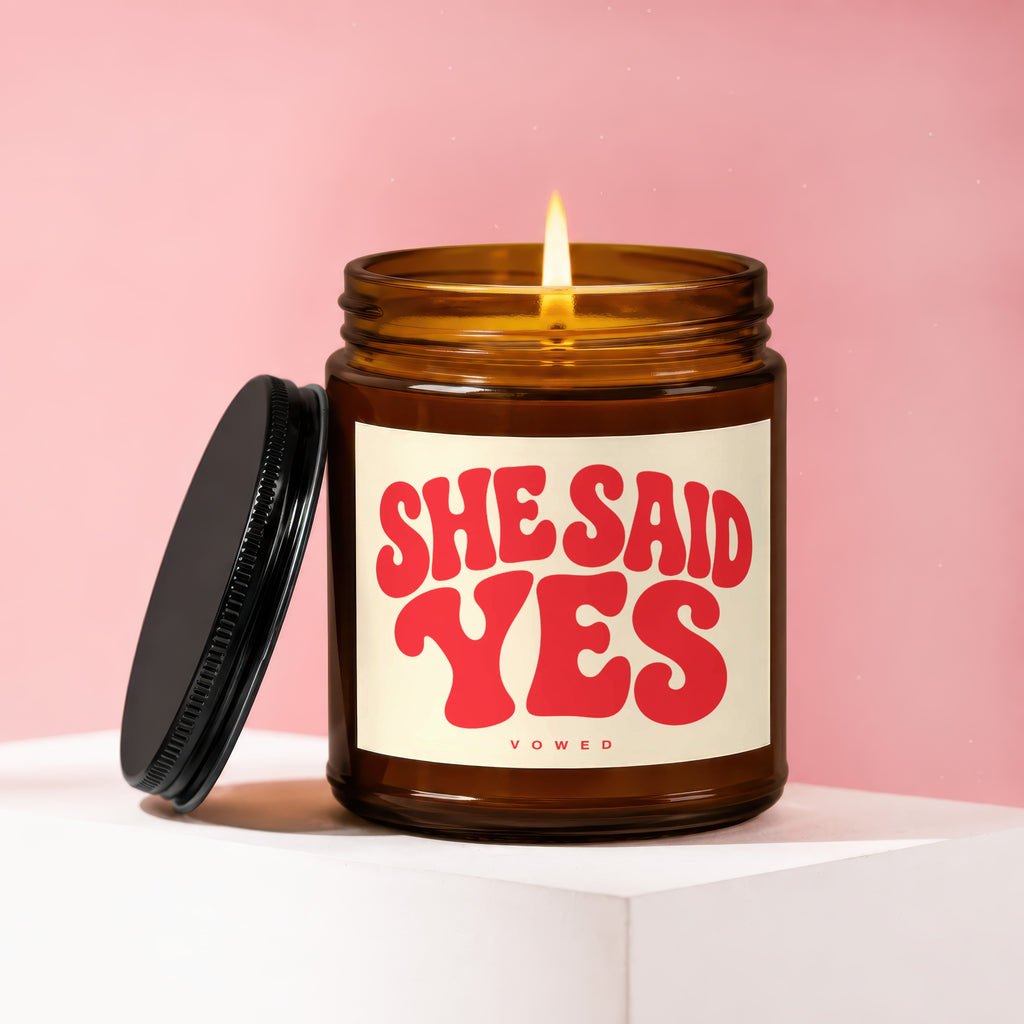 Amber candle with lit wick, lid off, labeled "SHE SAID YES VOWED".