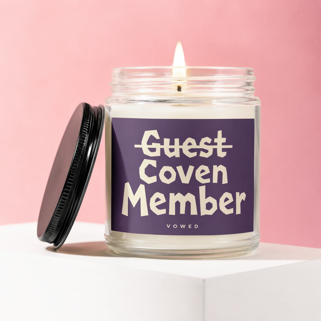 Glass jar candle with burning wick and a purple label reading "Guest Coven Member Vowed".