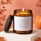 Glass candle with lit flame, black lid, and white label that reads "Keeping It Together... Mostly. VOWED".