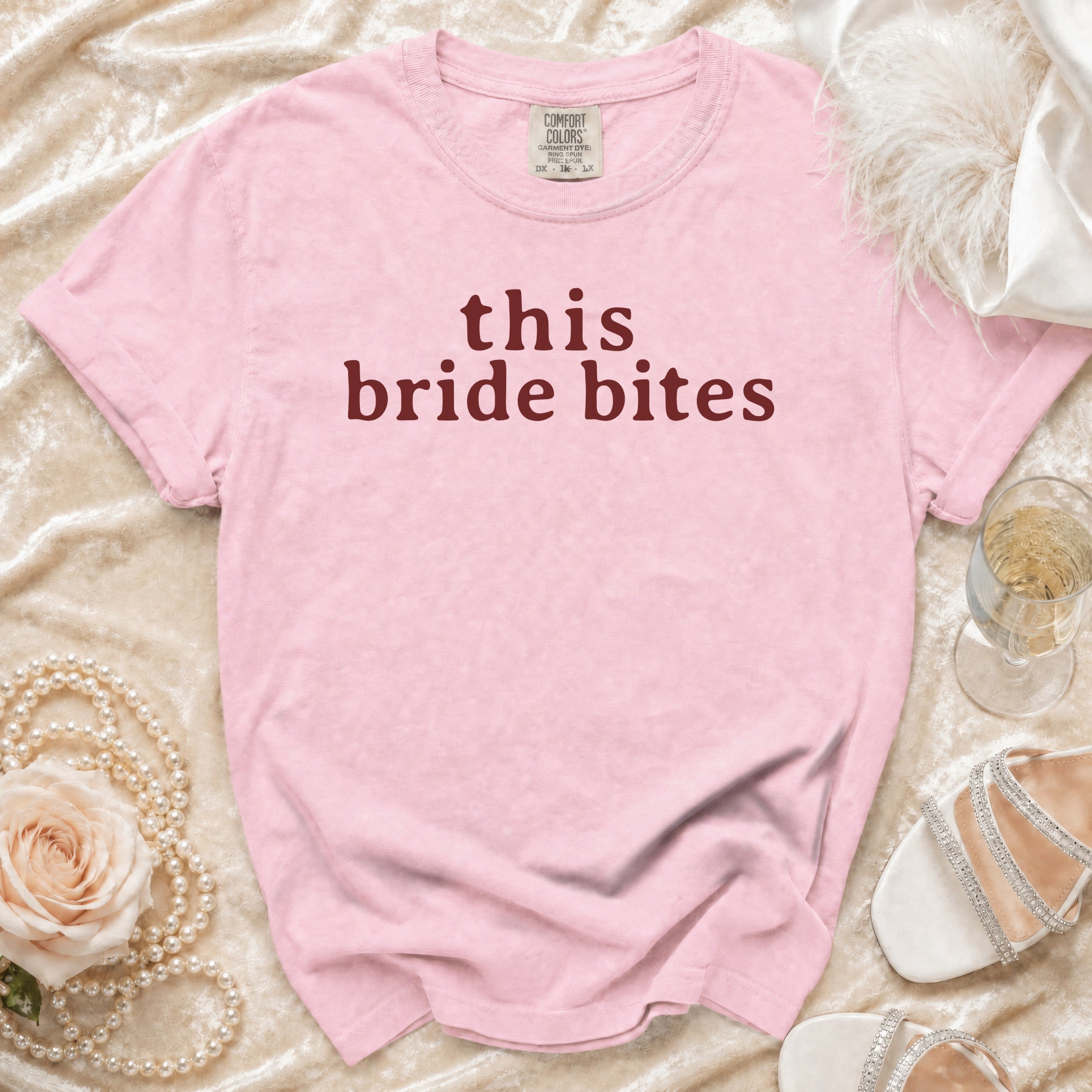 Pink t-shirt with "this bride bites" text, pearls, rose, and champagne.