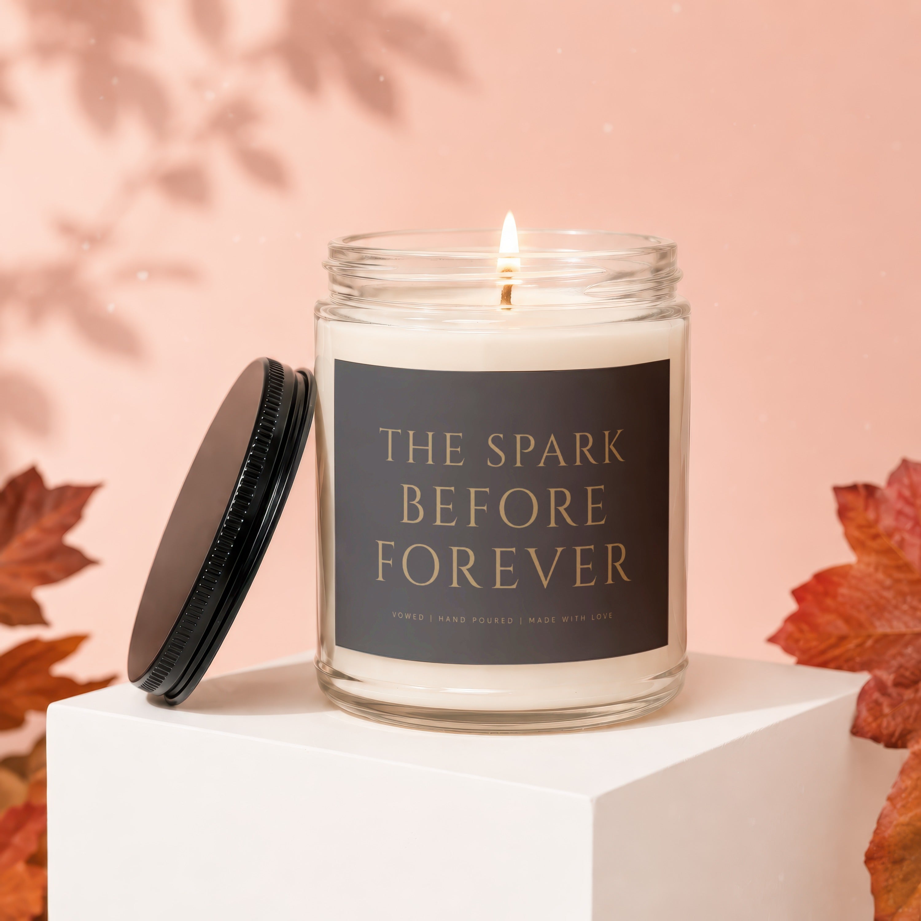 Candle with "The Spark Before Forever" text, lid off, surrounded by autumn leaves.