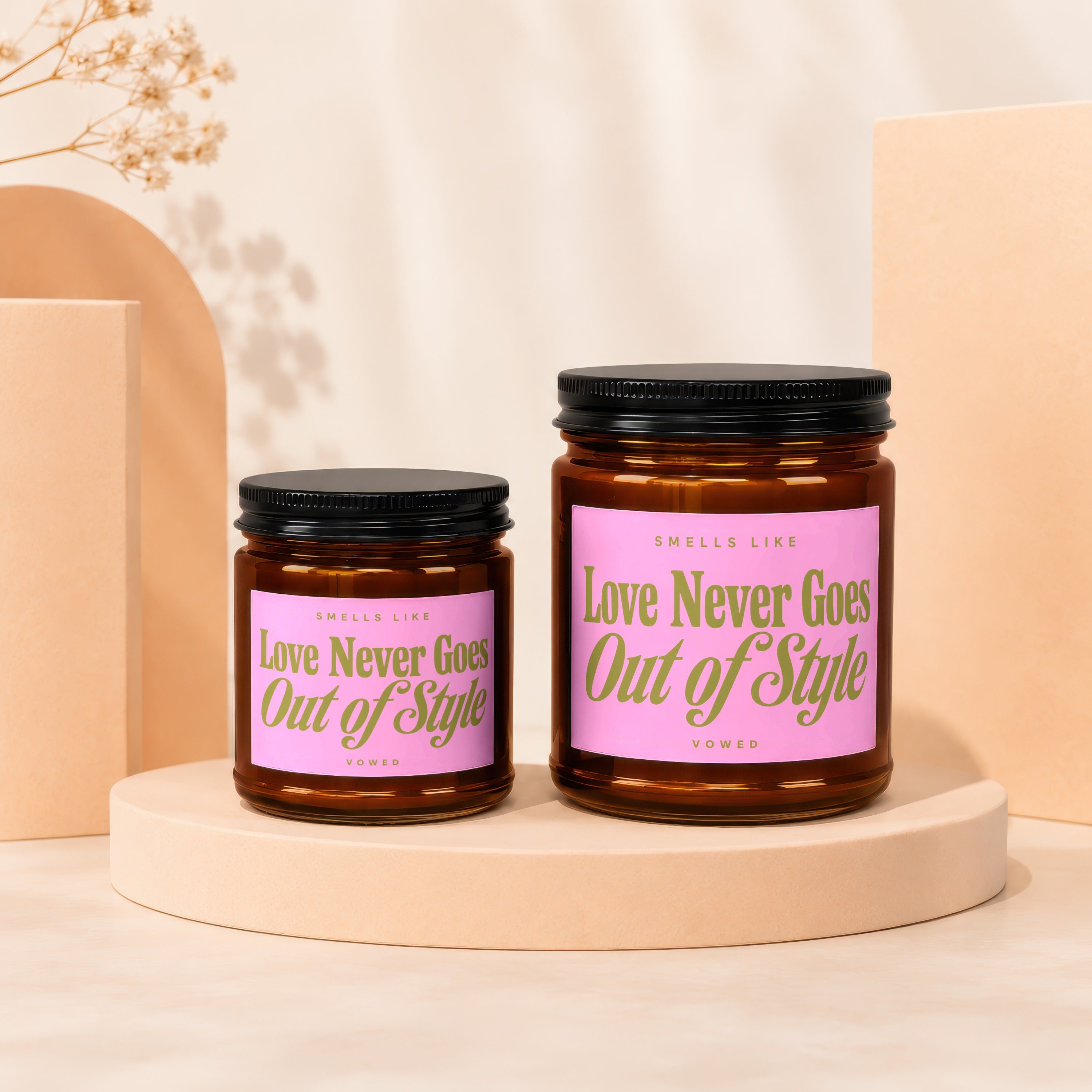 Two amber glass candles with pink labels that read "Love Never Goes Out of Style Vowed".
