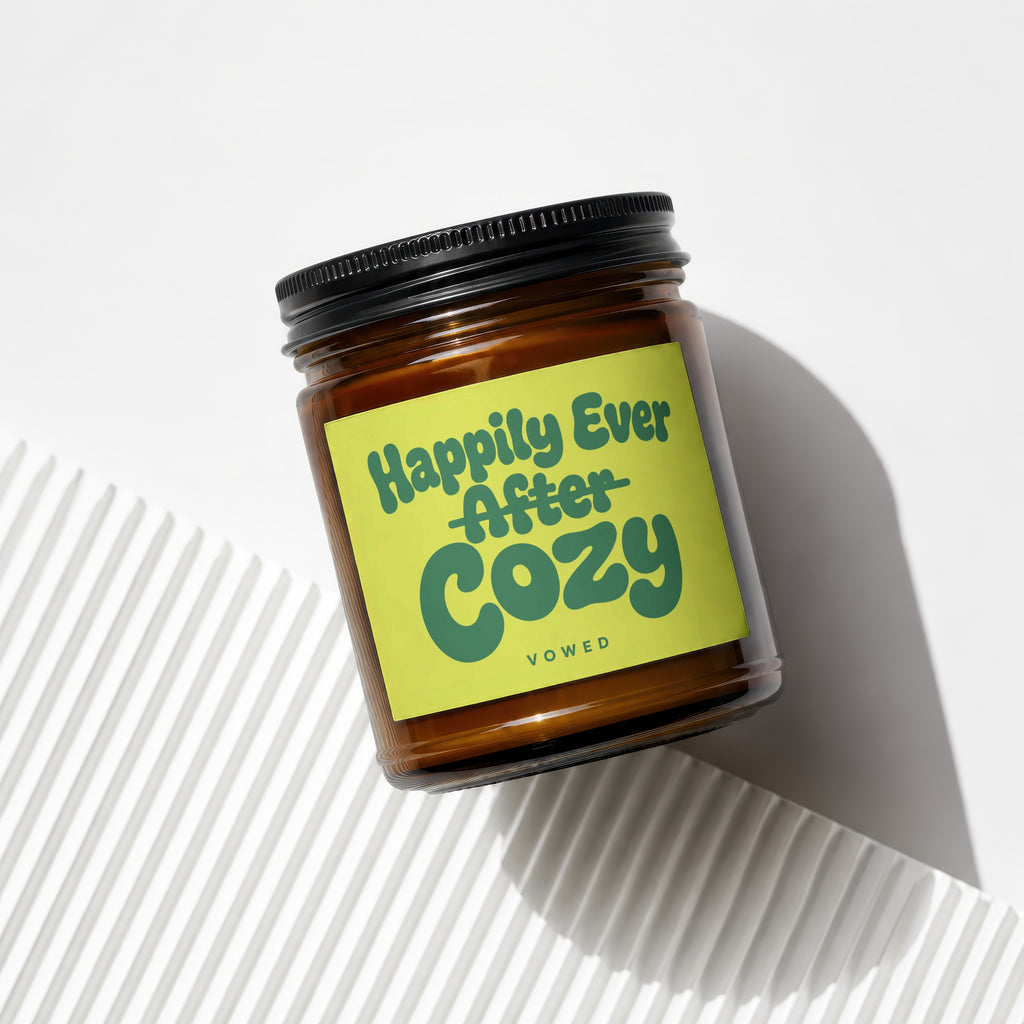 Amber glass candle with a neon green label reading "Happily Ever After Cozy Vowed".