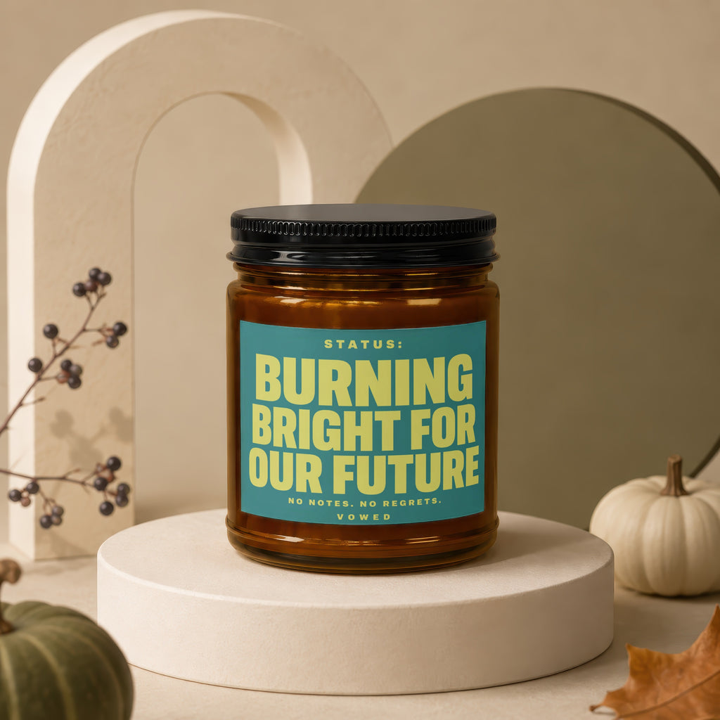Amber jar candle with teal label reading "STATUS: BURNING BRIGHT FOR OUR FUTURE".