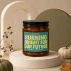 Amber jar candle with teal label reading "STATUS: BURNING BRIGHT FOR OUR FUTURE".