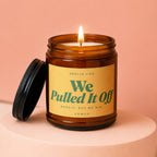 Amber jar candle with a lit flame and a black lid, labeled "We Pulled It Off"