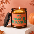 Lit amber glass candle with orange label reading "MY Hostages BRIDESMAIDS"