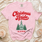 A pink Comfort Colors t-shirt with red text reading Christmas Bride above a green church graphic and the date 12.19.26.